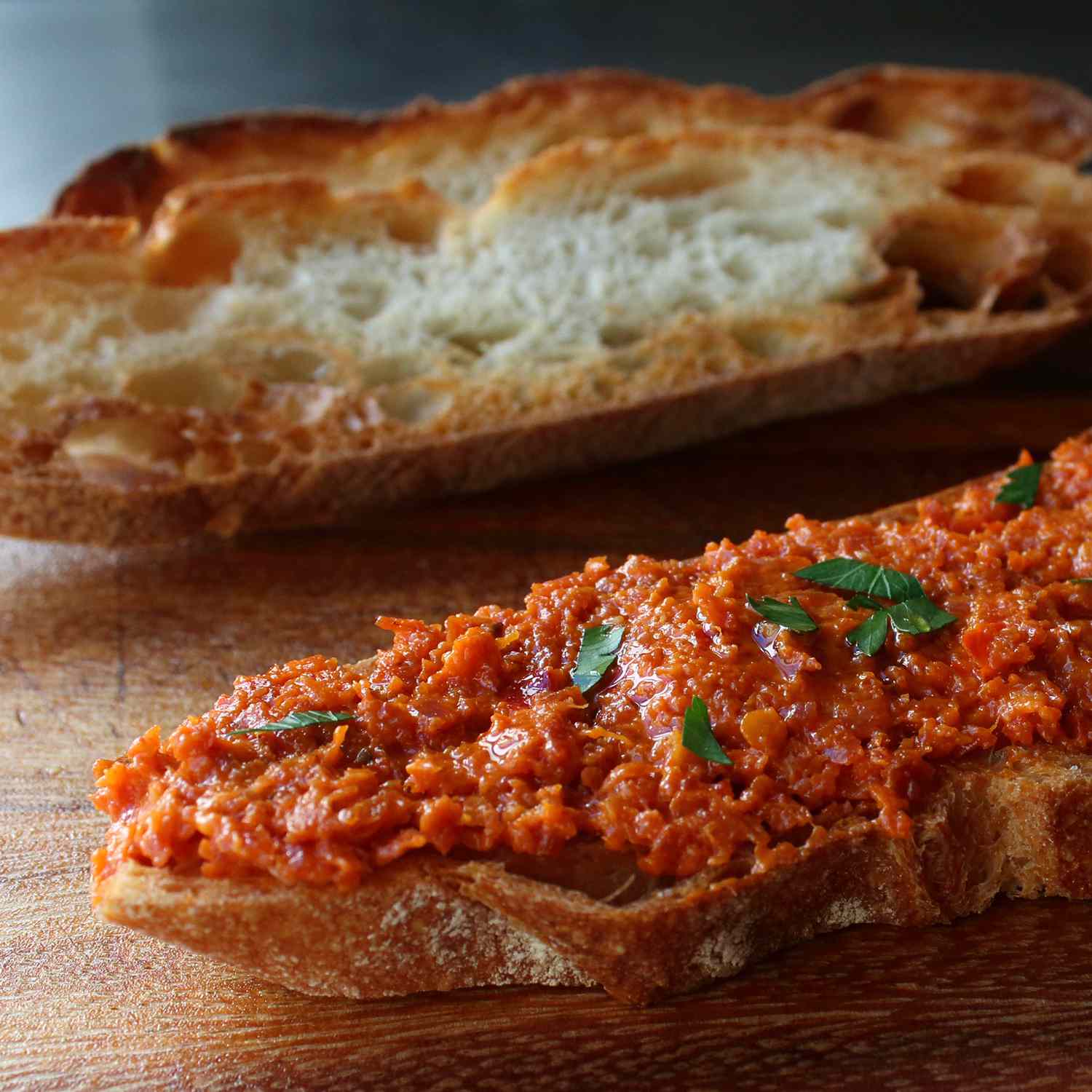 Close-up of 'nduja spread over toasted sourdough with olive oil and fresh parsley, a torn baguette behind it on a wooden board