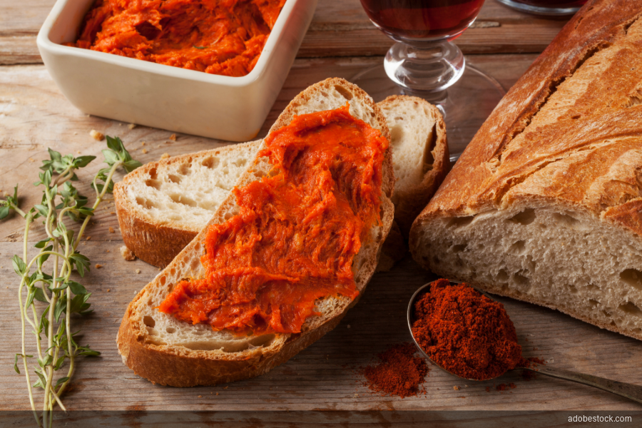 Spoonful of Calabrian 'nduja spread thickly across a slice of crusty bread, with a bowl of 'nduja, fresh thyme, ground chili and a glass of red wine on a rustic wooden table